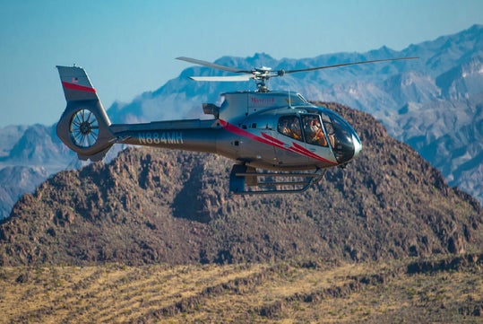 A silver and red helicopter with visible registration N884MH flies over a rocky desert landscape with mountains in the background.