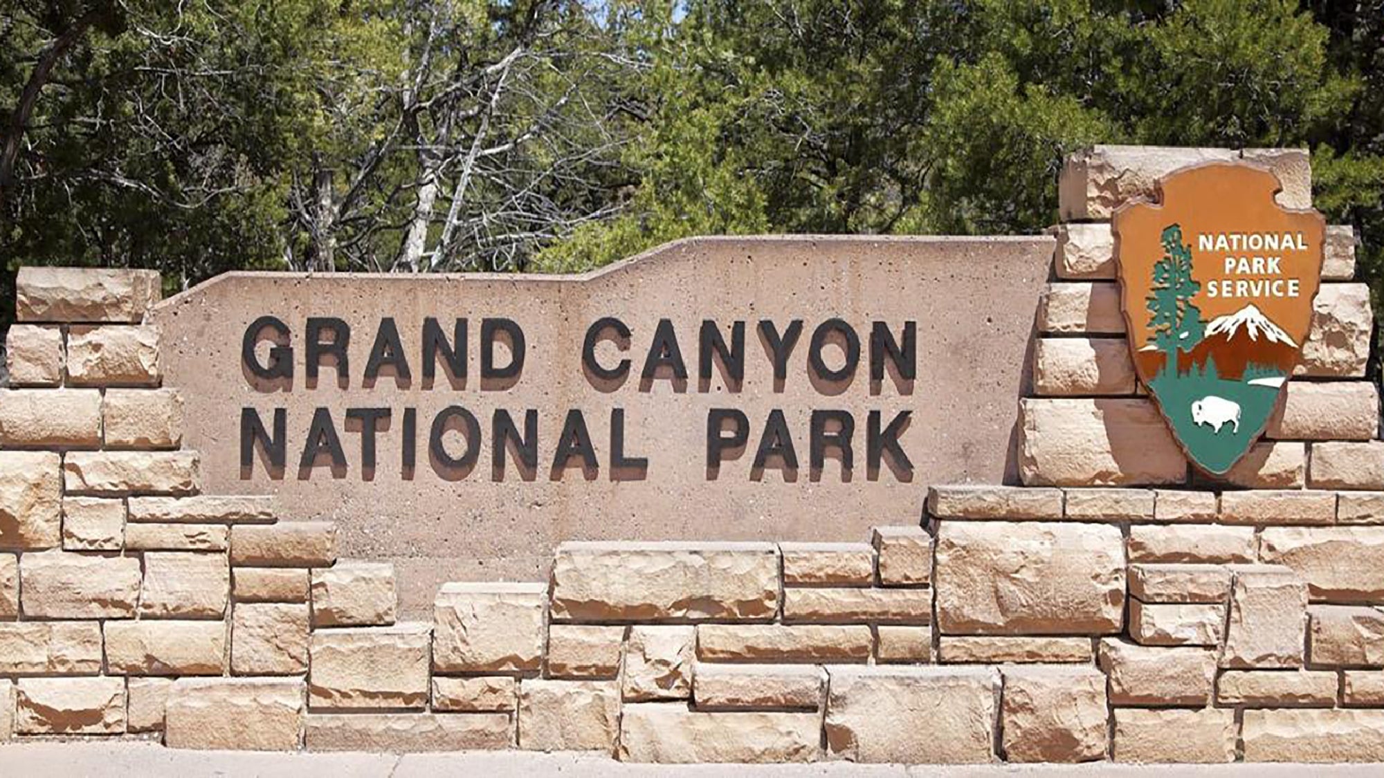 Stone entrance sign for Grand Canyon National Park with the National Park Service emblem on the right, surrounded by trees in the background.