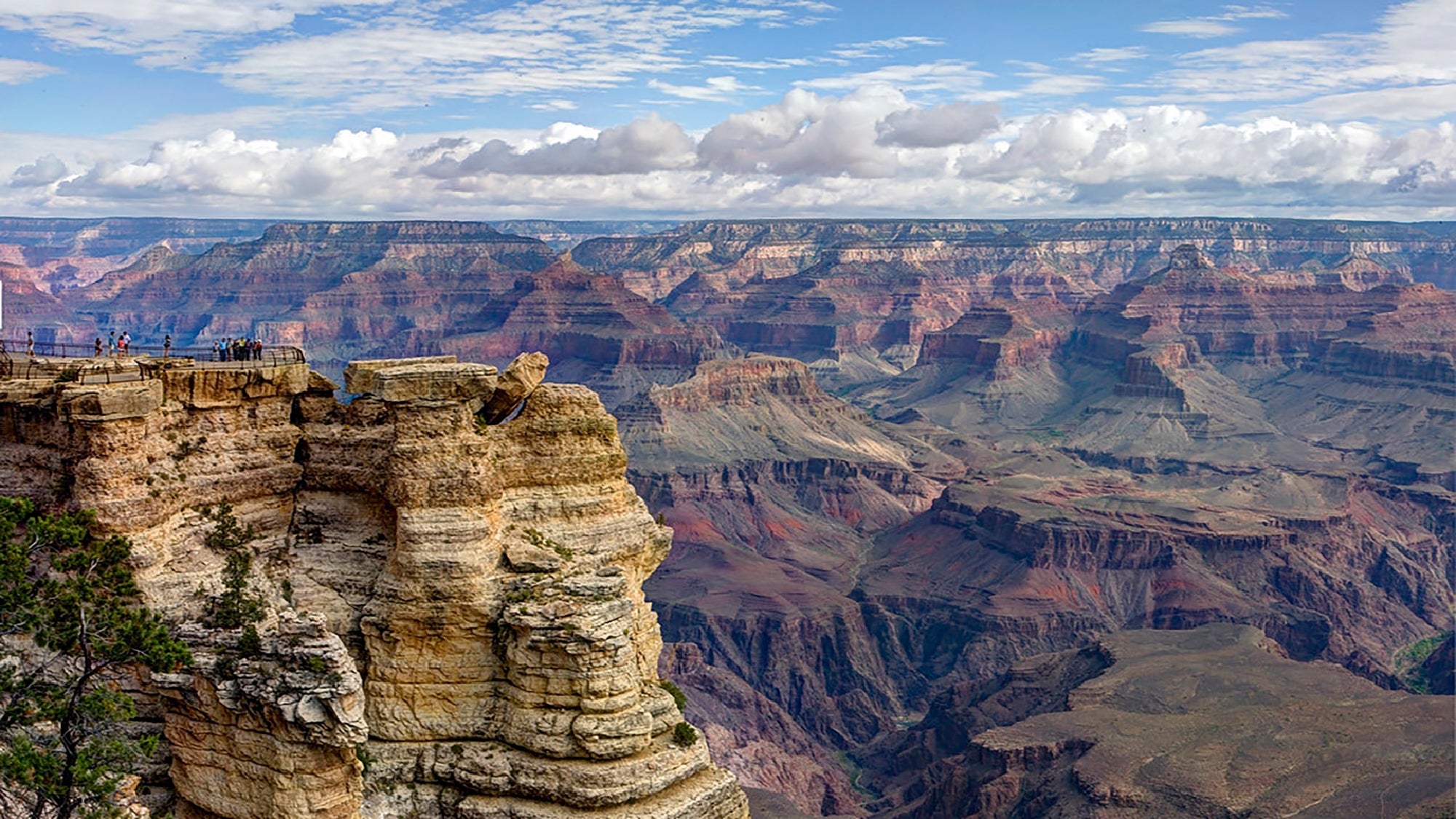 View of the Grand Canyon under a partly cloudy sky, with layered rock formations and tourists standing on a lookout point near the edge.