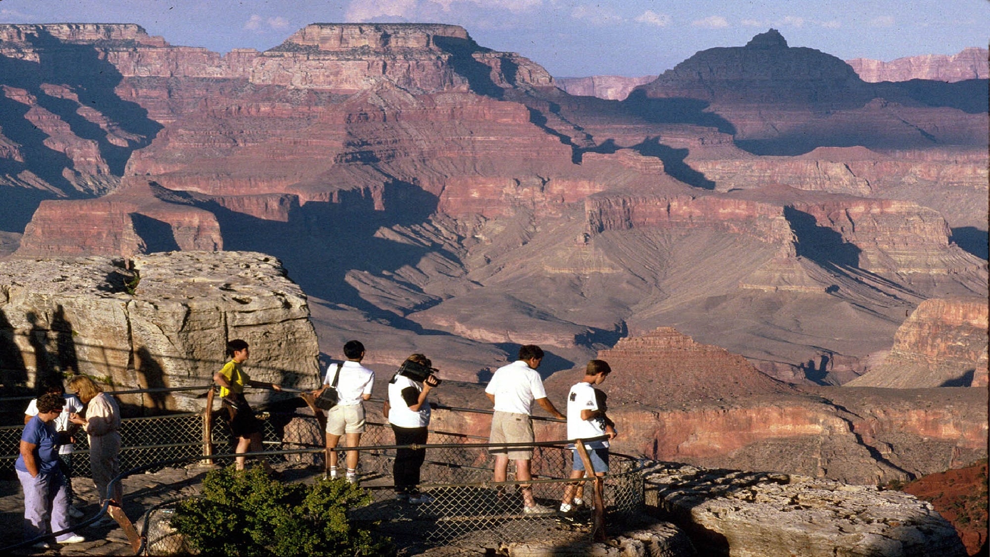 A group of people stands at a fenced viewpoint overlooking the layered rock formations of the Grand Canyon under a clear sky.