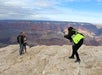 Person in a neon yellow jacket takes a photo of a couple standing on a rocky ledge with the Grand Canyon in the background under a partly cloudy sky.