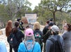 A group of people listen to a guide speaking in front of an informational sign outdoors, surrounded by trees and stone pathways.