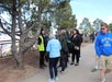 A group of people stands on a paved path near a fence and trees; one person in a bright vest appears to be speaking to the group.