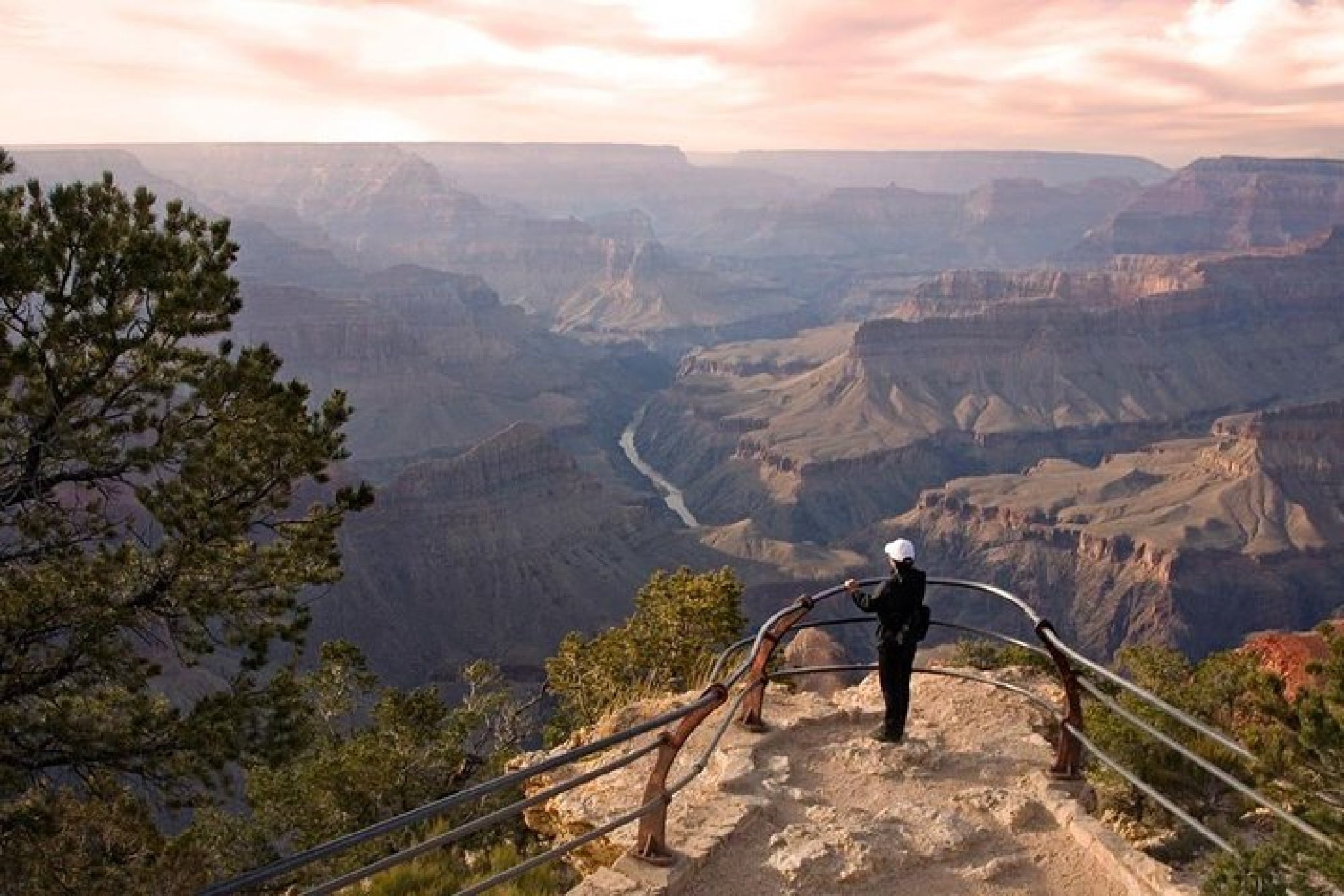 A person stands at a fenced viewpoint overlooking the Grand Canyon, with layered rock formations and the Colorado River visible below under a partly cloudy sky.