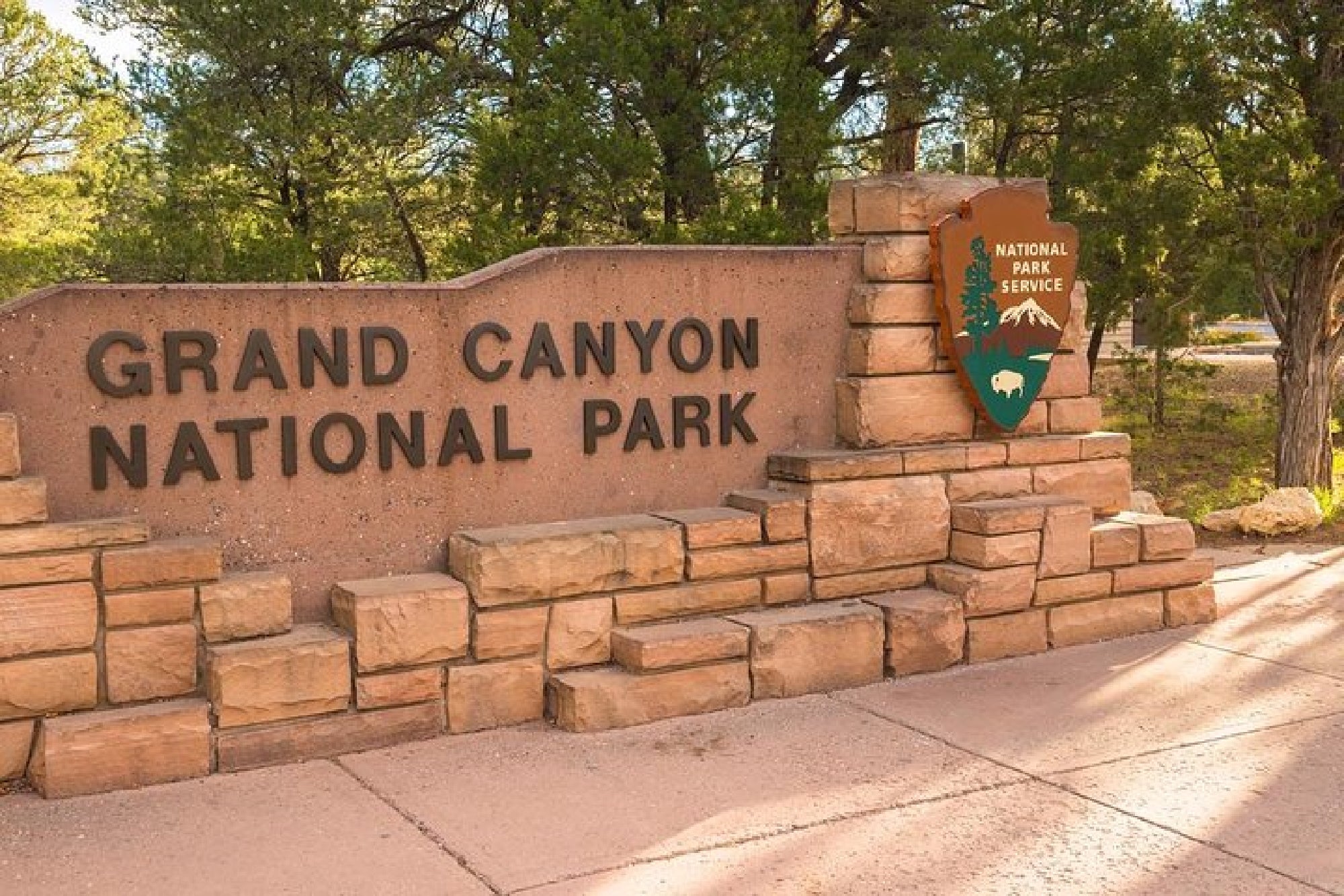 Entrance sign for Grand Canyon National Park, featuring stonework and the National Park Service emblem, surrounded by trees.