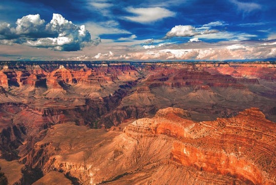 A wide view of the Grand Canyon shows layered red rock formations under a partly cloudy blue sky.