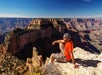 A man in an orange shirt and shorts sits on a rocky edge overlooking the Grand Canyon under a clear blue sky.