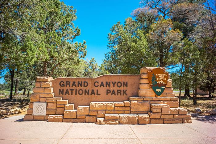 Stone entrance sign for Grand Canyon National Park, surrounded by trees and a National Park Service emblem on a sunny day.