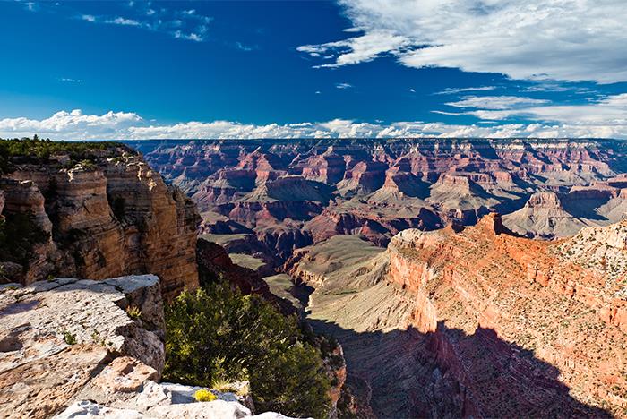 View of the Grand Canyon with layered rock formations, deep valleys, and a clear blue sky with scattered clouds.