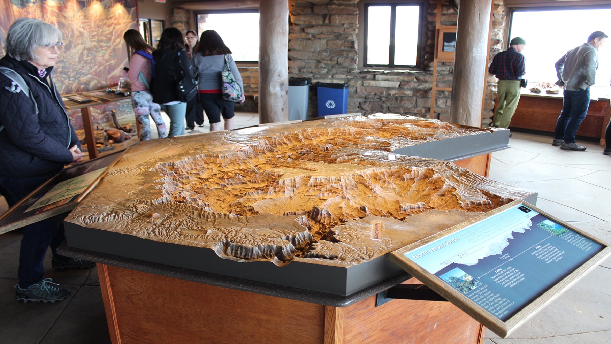 Visitors observe a detailed 3D topographic model of the Grand Canyon inside an indoor exhibit space with informational displays.