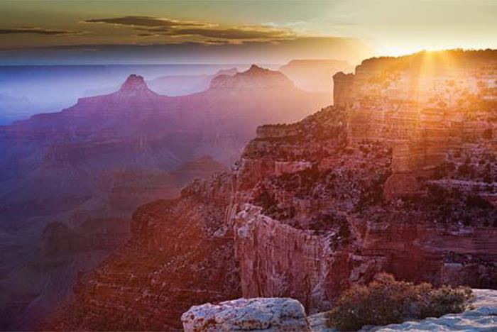 Sunrise over the Grand Canyon, with sunlight illuminating layered rock formations and casting shadows across the vast landscape.