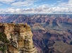 View of the Grand Canyon with layered rock formations, deep gorges, and a group of people standing on a rocky overlook under a partly cloudy sky.