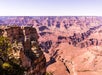 A wide view of the Grand Canyon with layered rock formations, deep valleys, and groups of people standing on an overlook near the edge.