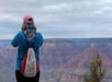 A person wearing a pink cap and blue jacket faces the Grand Canyon, holding up a phone to take a photo.