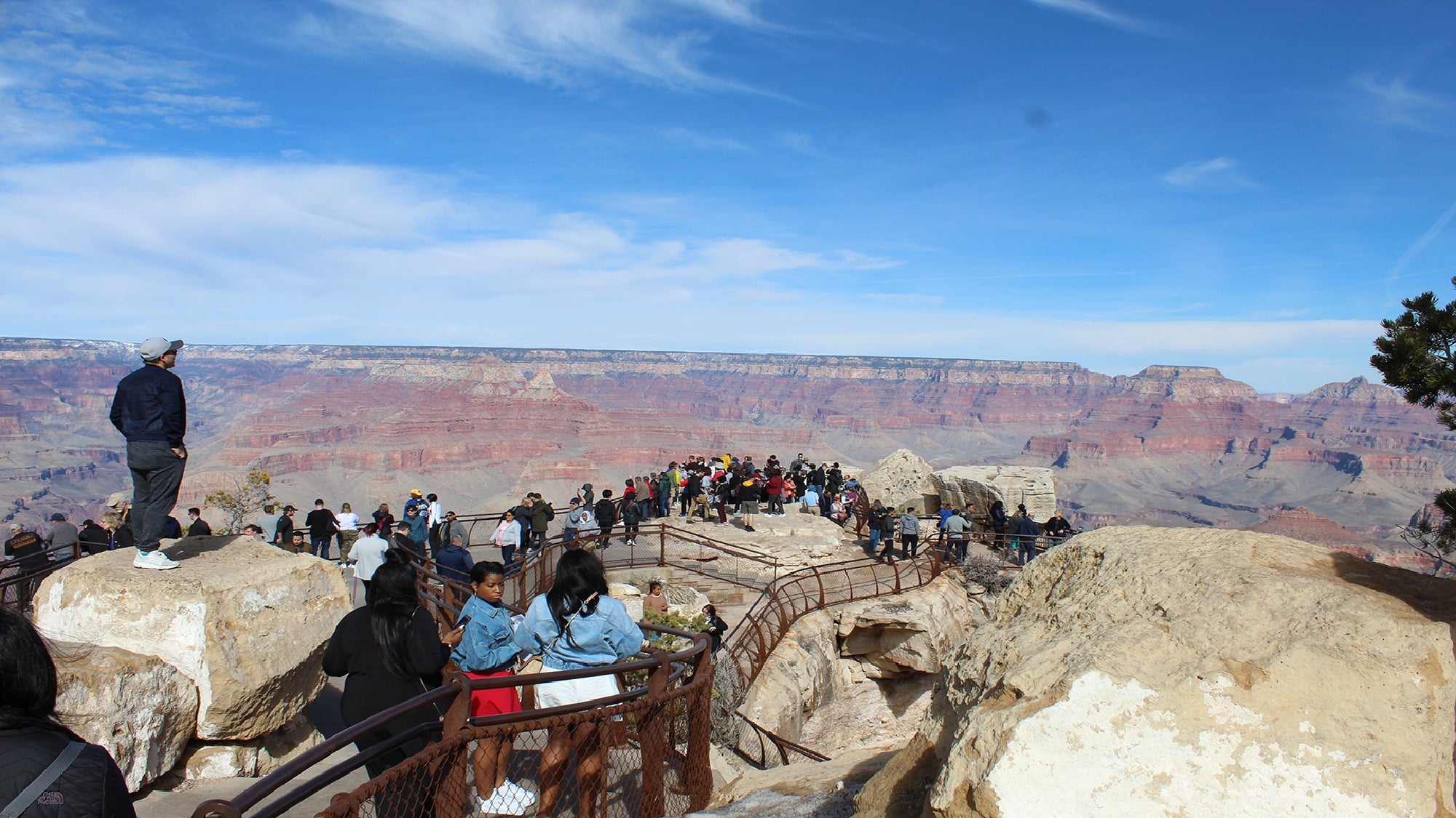 Crowds of people stand on a rocky overlook with railings, viewing the Grand Canyon’s layered cliffs and vast landscape under a blue sky.