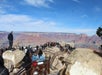 Crowds of people stand on a rocky overlook with railings, viewing the Grand Canyon’s layered cliffs and vast landscape under a blue sky.