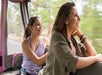 Two women sit together in a moving vehicle, looking out the open window at blurred greenery, both appearing relaxed and happy.