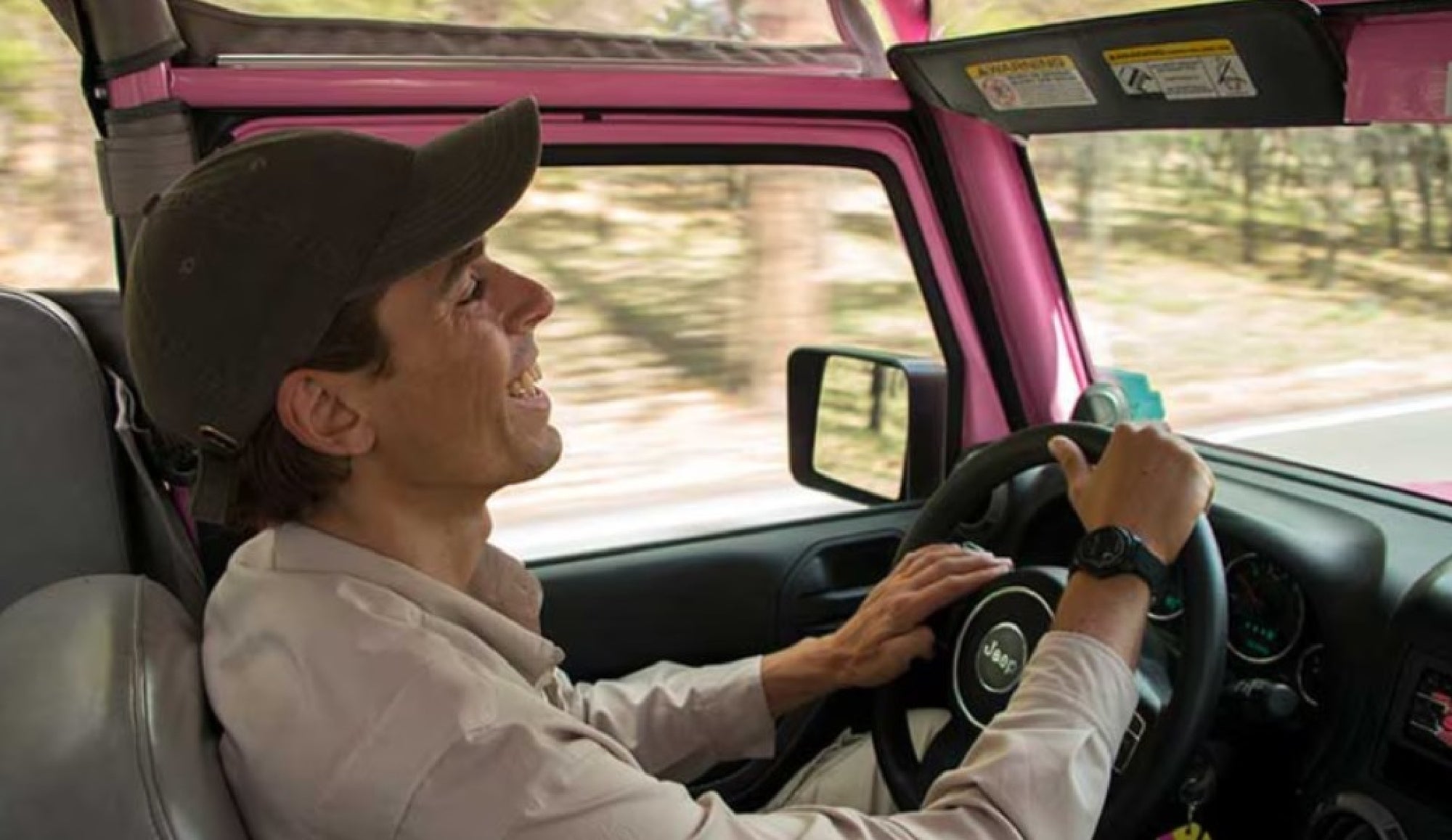 A man wearing a cap drives a pink Jeep with the roof open on a forested road during the day.
