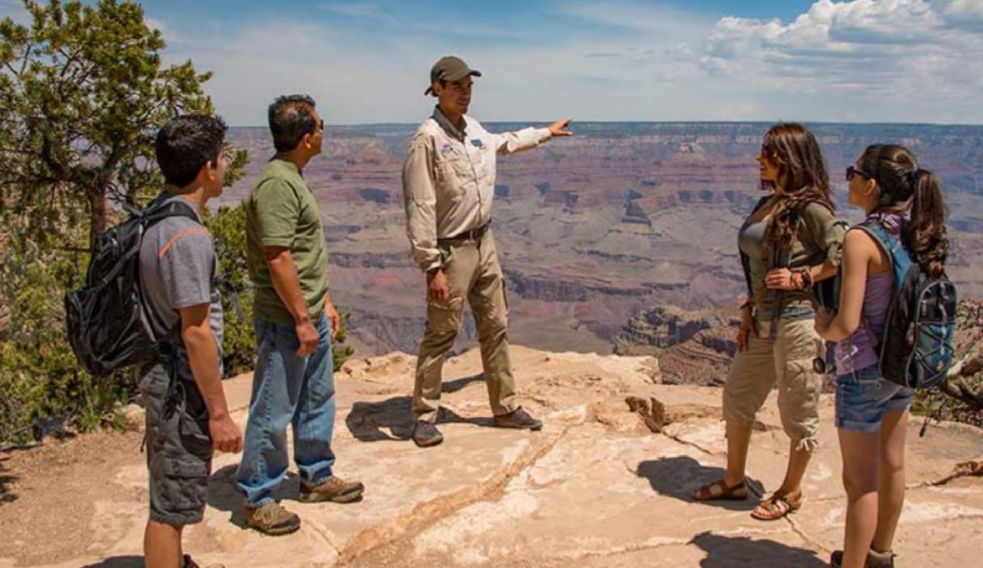 A tour guide speaks to four visitors standing at the edge of a canyon, with rocky cliffs and a wide canyon landscape in the background.