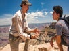 A park ranger talks to a teenage boy near the edge of a canyon under a blue sky, with rocky cliffs and desert landscape in the background.