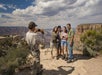 A person takes a photo of four people posing together on a rocky overlook with the Grand Canyon in the background under a partly cloudy sky.