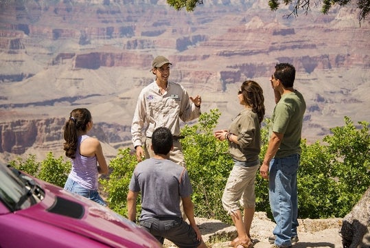 A park ranger speaks to a group of four visitors by a railing overlooking the Grand Canyon on a sunny day.
