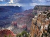 View of the Grand Canyon’s layered red rock formations under a cloudy sky, with distant rain visible on the horizon.