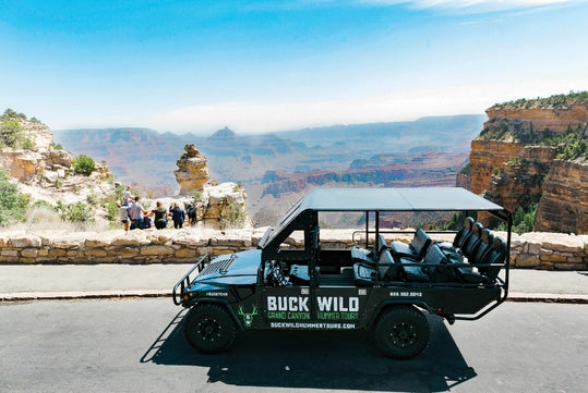 An open tour vehicle labeled "Buck Wild Grand Canyon Hummer Tours" is parked at a scenic Grand Canyon overlook with tourists and canyon views in the background.