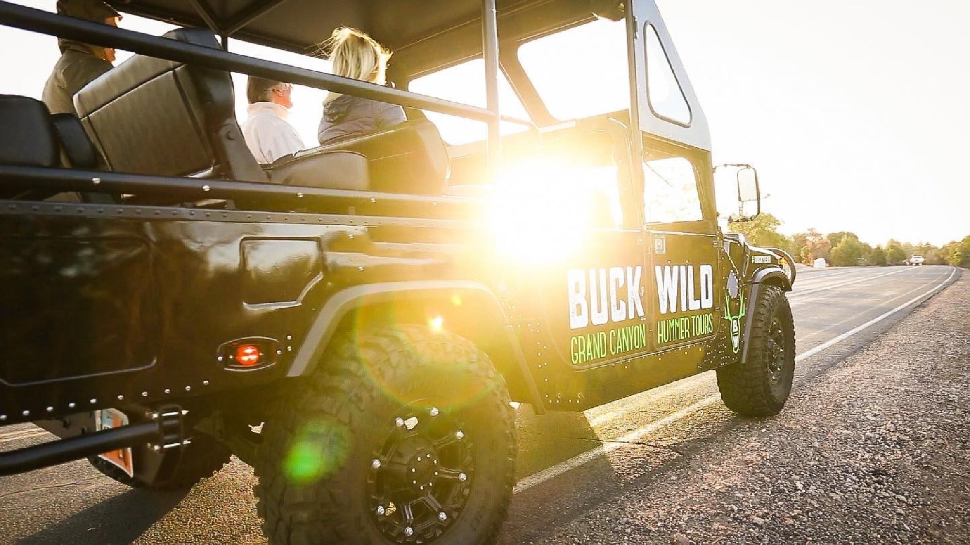 Tour vehicle with "Buck Wild Grand Canyon Hummer Tours" on the side, carrying passengers on a road at sunset with sunlight shining through the windows.