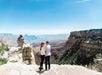 A couple stands together on a rocky overlook at the Grand Canyon, with expansive canyon views and clear blue sky in the background.