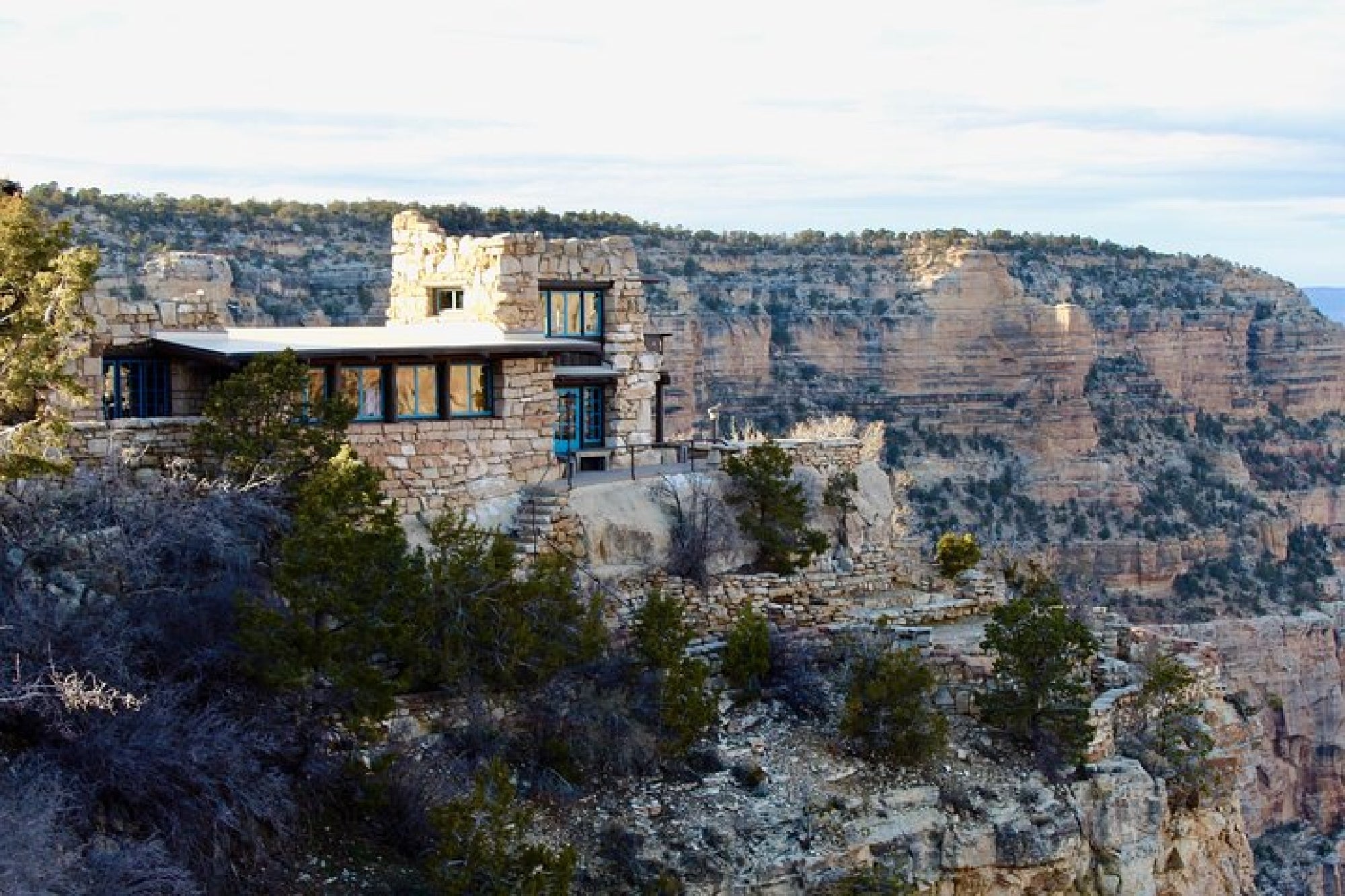 A stone building with large windows sits on the edge of a cliff, overlooking a canyon landscape with rocky walls and sparse vegetation.