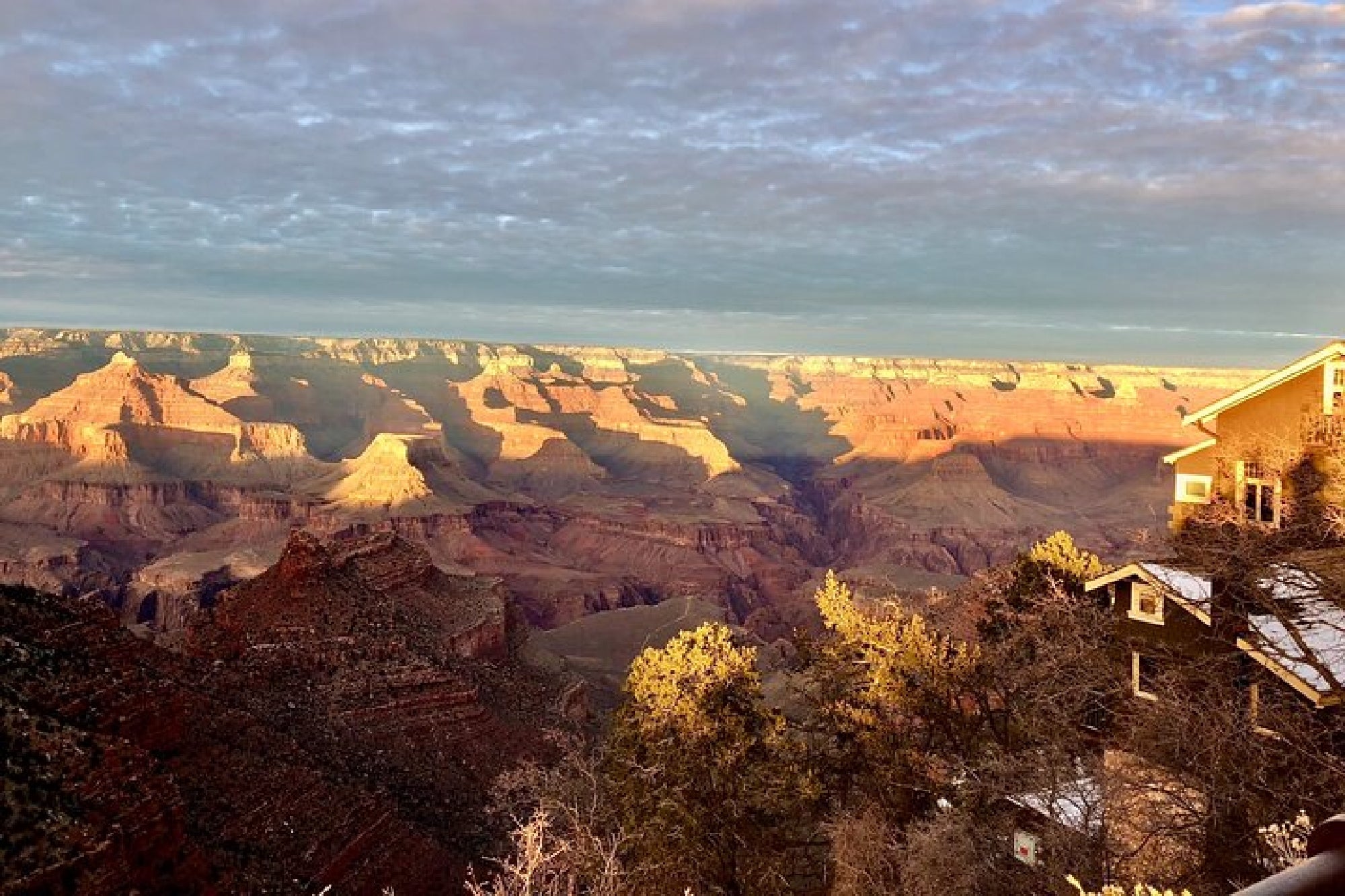Grand Canyon landscape at sunset with layered rock formations and shadows; a building is partially visible on the right side amid trees.