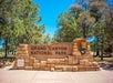 Entrance sign for Grand Canyon National Park, made of stone, with trees and a National Park Service emblem in the background.
