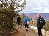 Three people with backpacks walk on a paved trail near the edge of the Grand Canyon, with trees and canyon views in the background.