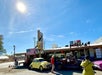 A roadside diner on Route 66 features vintage cars, a large Route 66 sign, and an American flag under a bright, sunny sky with people outside.