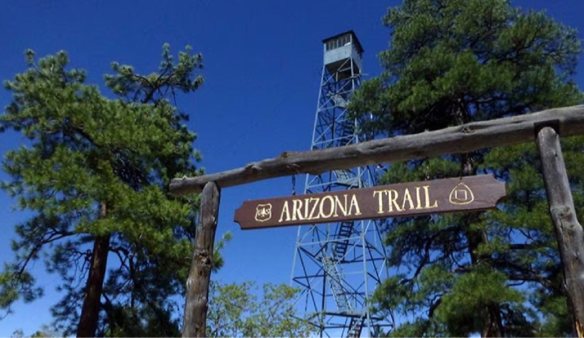 A wooden sign reading "Arizona Trail" stands in front of pine trees and a tall metal watchtower under a clear blue sky.