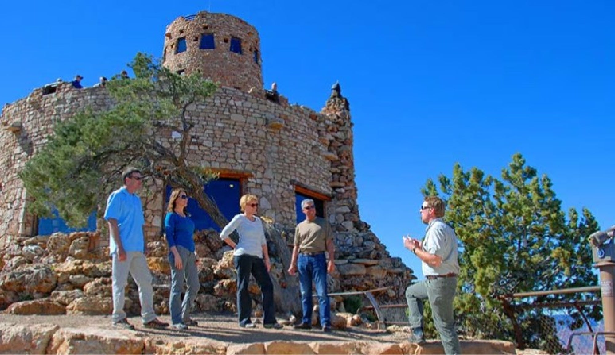A group of people stand near a stone lookout tower, listening to a guide speak, with clear blue sky and trees in the background.