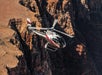 A helicopter with red and white markings flies above rocky canyon cliffs during the daytime.
