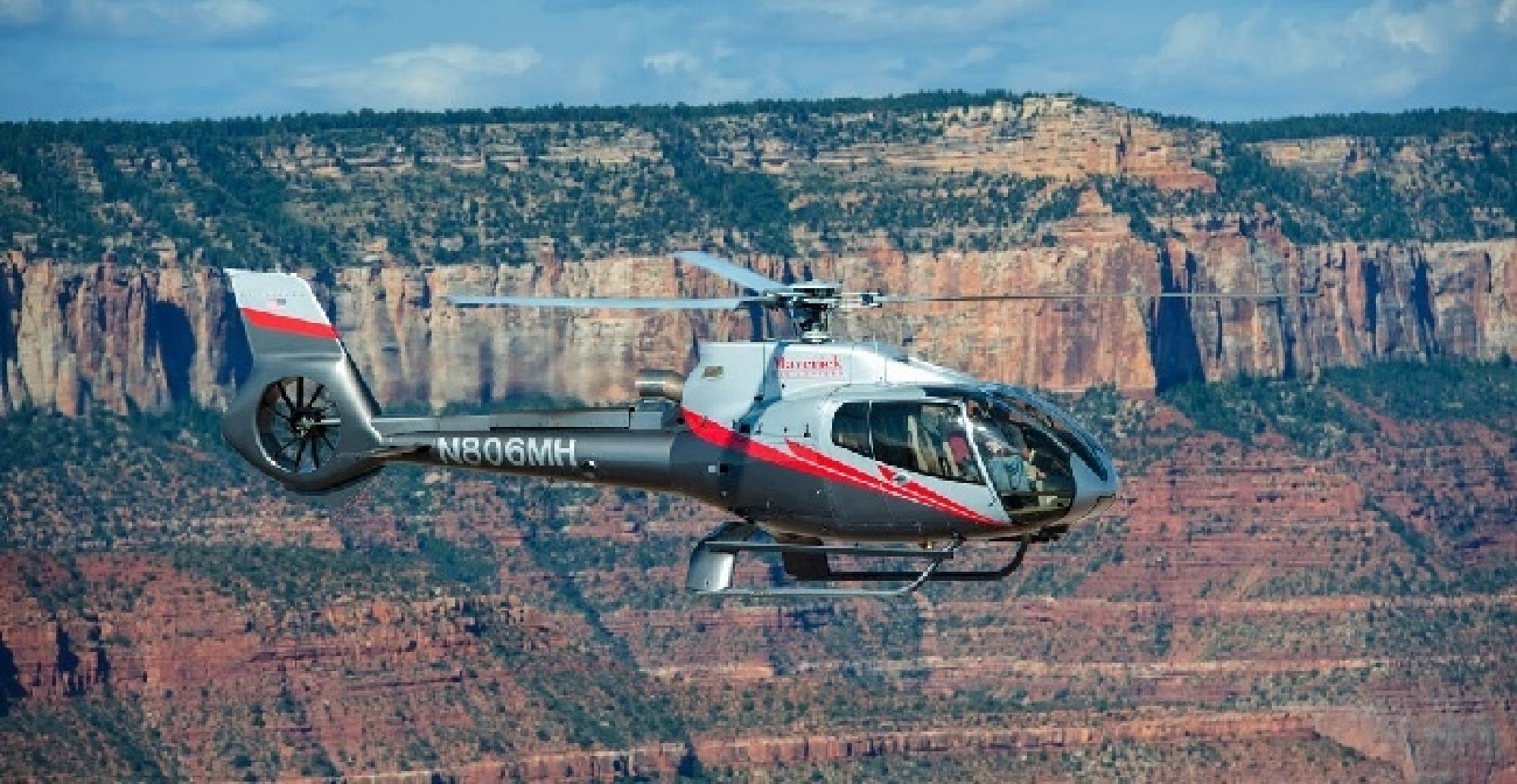 A gray helicopter with red accents flies in front of layered red rock cliffs under a blue sky.