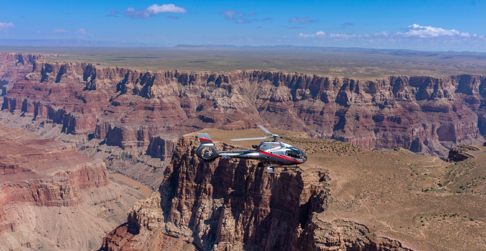 A helicopter flies over the Grand Canyon, with layered red rock formations and the Colorado River visible below under a clear sky.