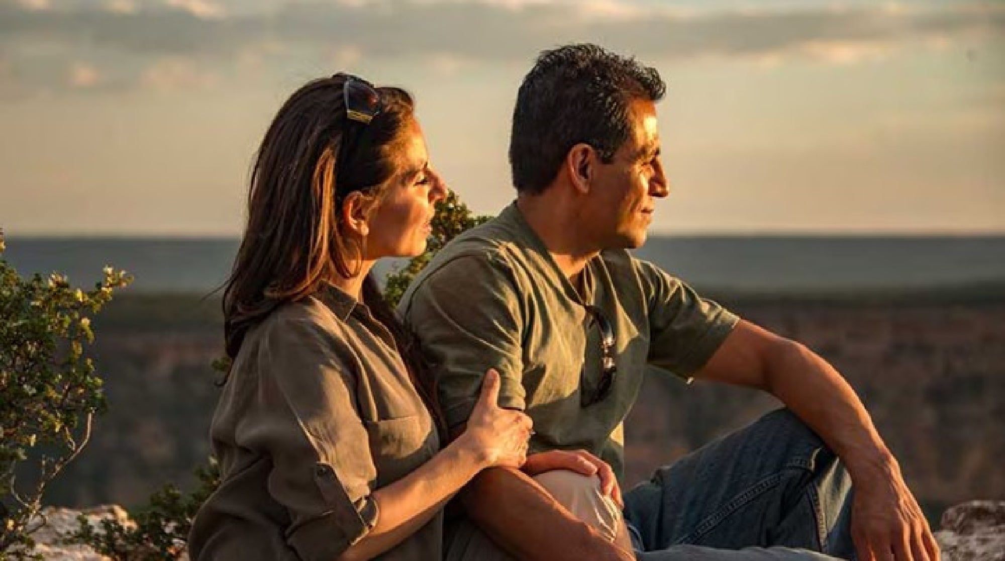 A man and woman sit close together outdoors at sunset, looking into the distance with relaxed expressions.