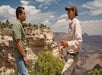 Two men stand and talk near the edge of a rocky cliff with canyon views and vegetation under a partly cloudy sky.