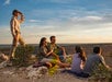 Four people sit on a rocky ledge with a scenic view, while one person stands and looks into the distance under a blue sky.