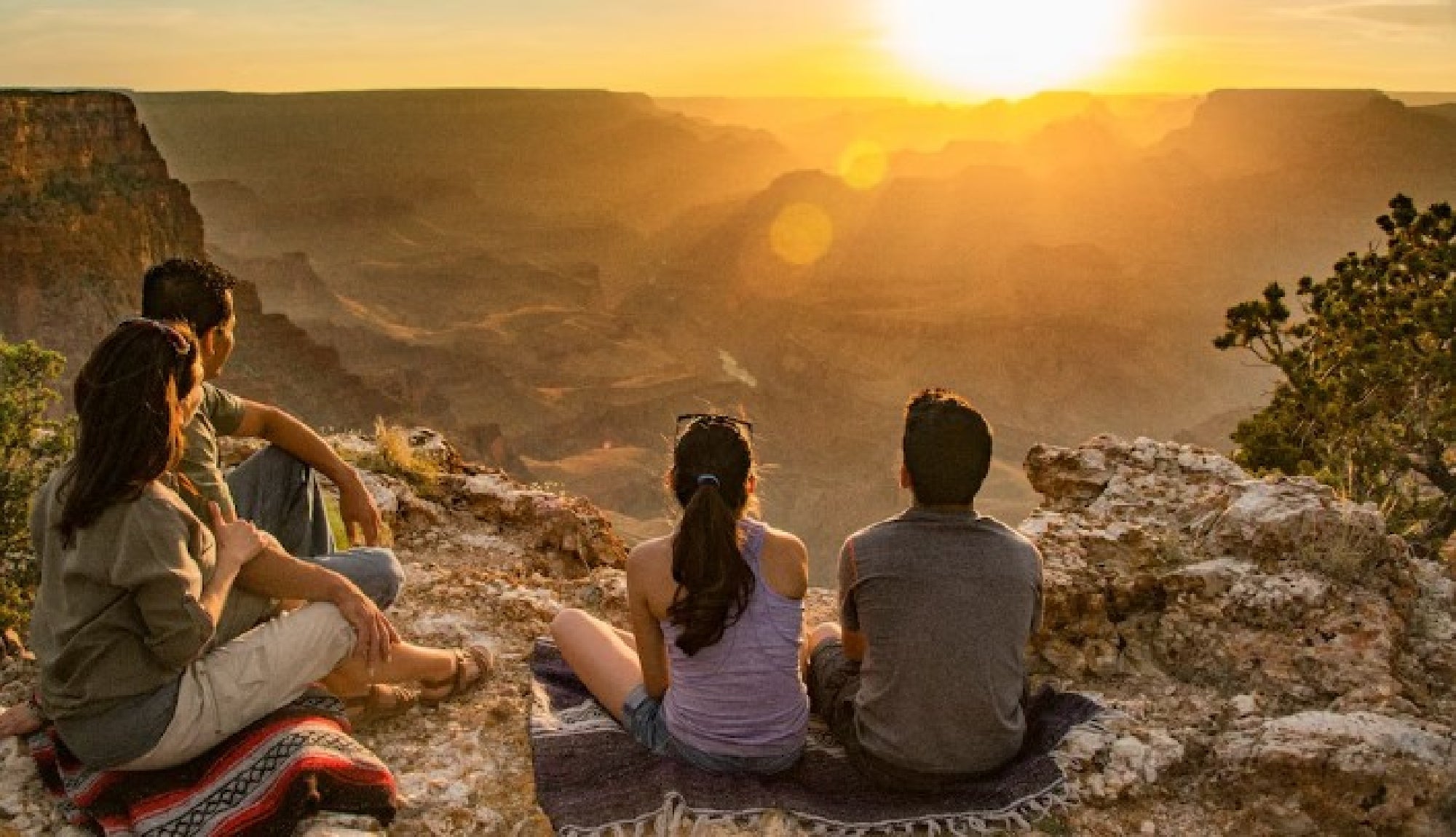 Four people sit on blankets near the edge of a rocky cliff, watching the sun set over a vast canyon landscape.