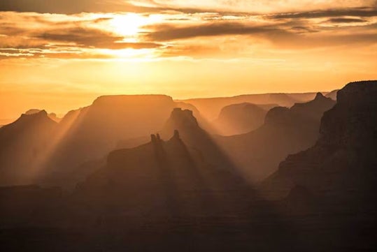 Sunlight streams through clouds over rugged canyon formations, casting long shadows and highlighting the landscape’s layers at sunrise or sunset.