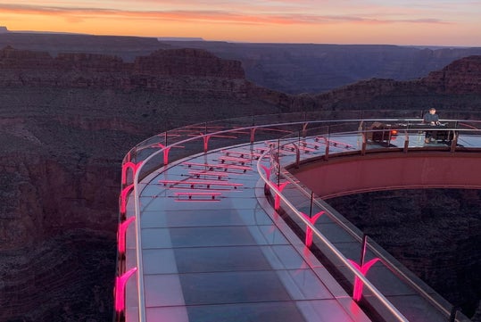 A curved glass skywalk with pink lights extends over a canyon at sunset; a person stands near the railing in the distance.