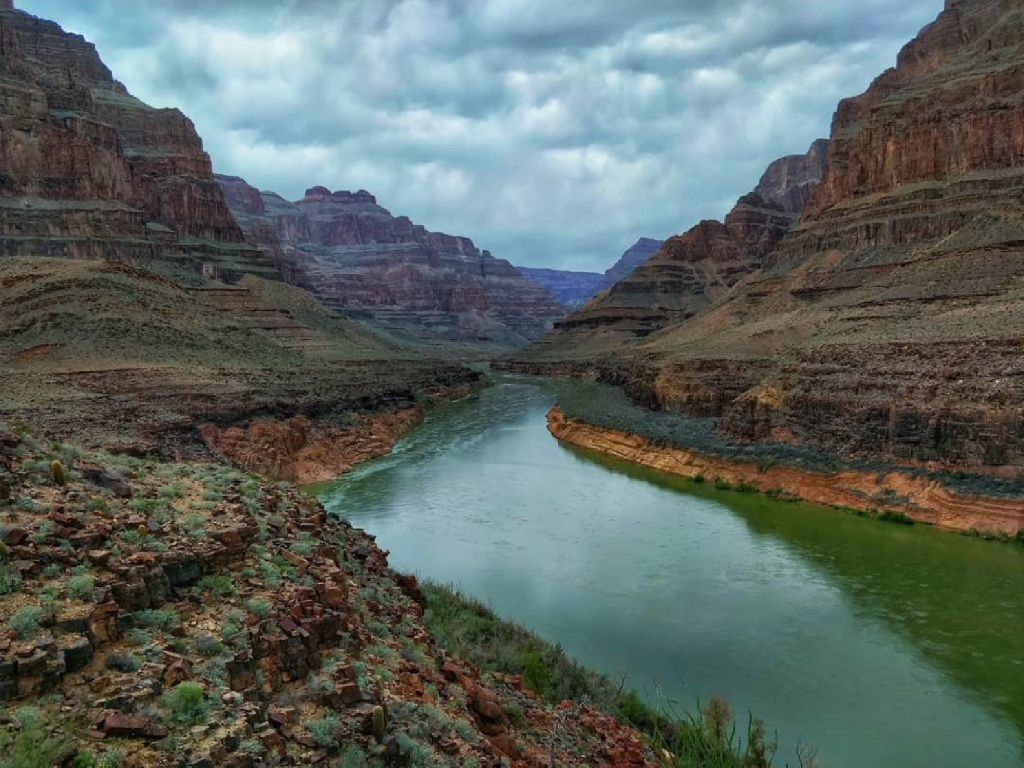 A wide river flows between steep, rocky canyon walls under a cloudy sky, surrounded by layered rock formations and sparse vegetation.