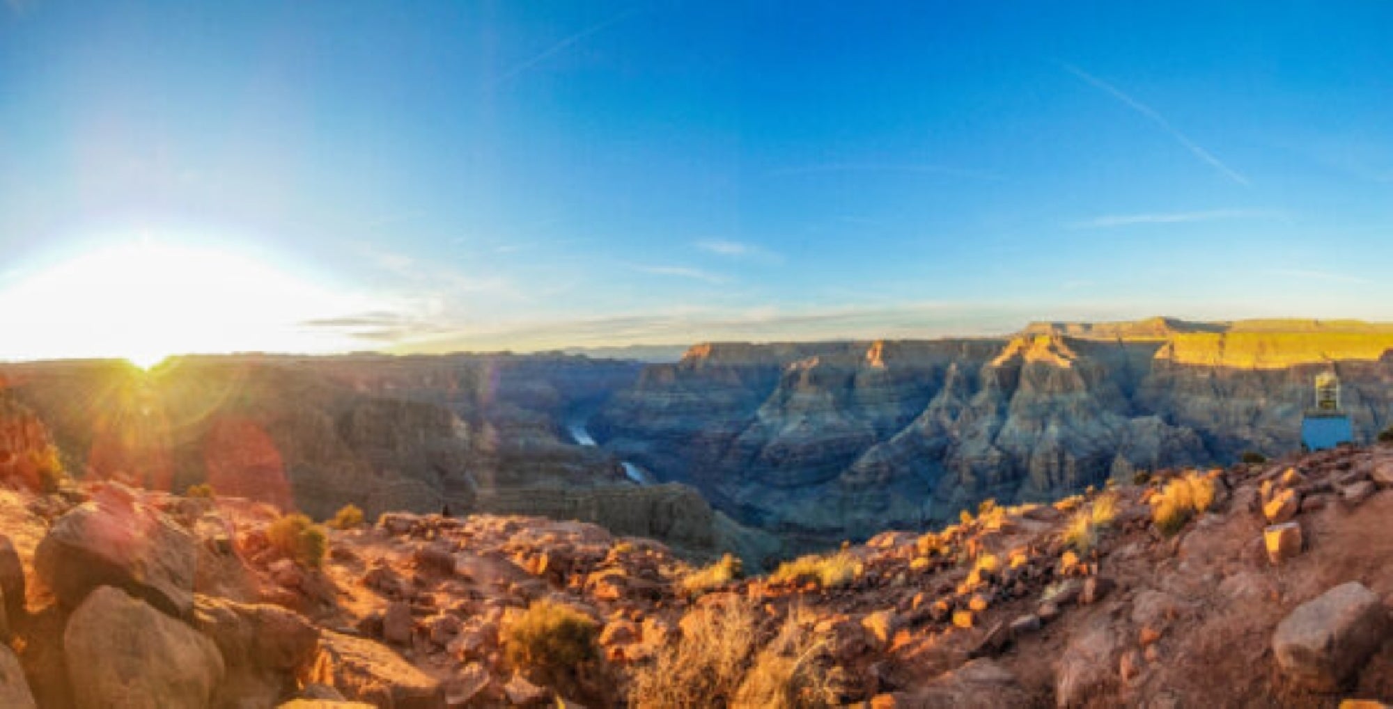 Panoramic view of the Grand Canyon at sunrise, showing rocky terrain, cliffs, and a river winding through the canyon under a clear blue sky.