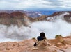 A person in a tan coat sits on a rocky ledge overlooking a cloudy, expansive canyon landscape under a partly cloudy sky.