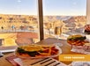 Two trays with burgers and fries on a table by a window overlooking the Grand Canyon Skywalk, with people walking outside. A drink and a "FOOD VOUCHER" sign are also visible.